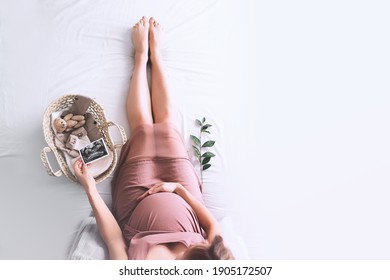 Pregnant woman in dress with ultrasound image. Mother with wicker basket of cute tiny stuff and teddy bear toy for newborn. Expectant mother waiting and preparing for baby birth during pregnancy. - Shutterstock ID 1905172507
