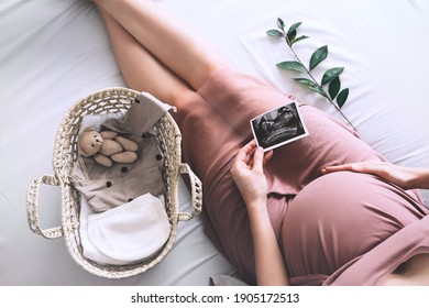 Pregnant woman in dress with ultrasound image. Mother with wicker basket of cute tiny stuff and teddy bear toy for newborn. Expectant mother waiting and preparing for baby birth during pregnancy. - Shutterstock ID 1905172513