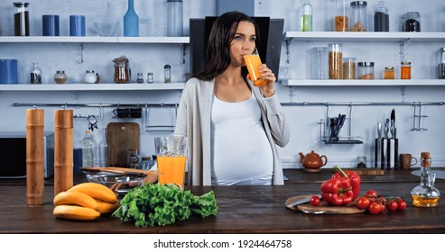 Pregnant woman drinking orange juice near organic food in kitchen