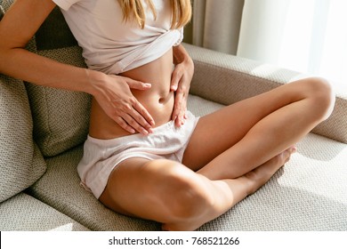 Pregnant Woman at an early pregnancy holding hands on belly sitting on sofa at home.