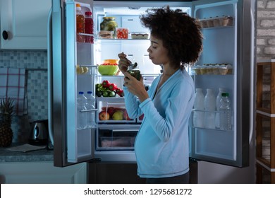 Pregnant Woman Eating Pickle From Jar Standing In Front Of An Open Refrigerator - Powered by Shutterstock