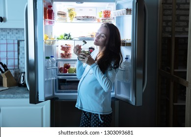 Pregnant Woman Eating Pickle From Jar In Front Of An Open Refrigerator