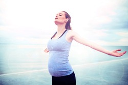 Pregnant Woman Embracing The Sunshine Standing Overlooking The Ocean With Her Arms Outstretched And Head Raised To The Sun With Closed Eyes