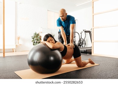 A pregnant woman is engaged in prenatal exercise on a fitness ball, supported by her partner in a bright studio setting. They focus on maintaining comfort and health during pregnancy. - Powered by Shutterstock