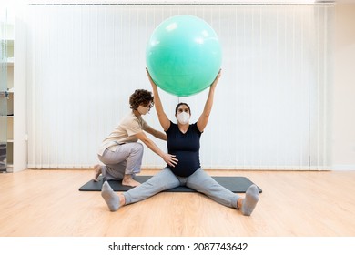 A pregnant woman with face mask sitting on a mat doing pilates exercises with a ball with the help of physiotherapist
