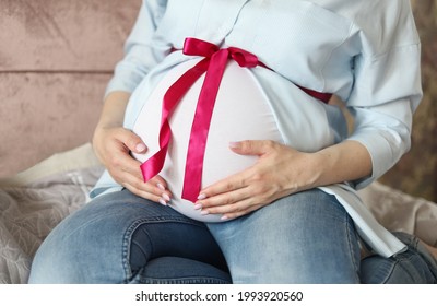 A pregnant woman. A woman gently wraps her arms around her pregnant tummy, with a pink ribbon and bow. Selective focus.