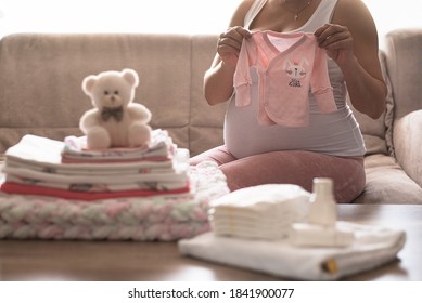 Pregnant Woman Is Getting Ready For The Maternity Hospital, Packing Baby Stuff. Pregnant Woman Preparing And Planning Baby Clothes. Pregnant Woman Holding Bodysuit New Born Baby. Preparing Stuff For
