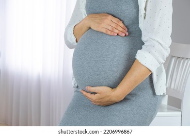 Pregnant Woman In Grey Dress And White Shirt Hold Her Big Tummy With Hands Near Window. Pregnancy, Maternity, Preparing And Expectation Concept. Baby Expectation, Care And Wellness On Pregnancy Time.