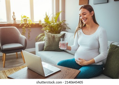 The pregnant woman is happy as she greets her family and friends on her laptop. She holds a glass of water while sitting on the sofa. The concept of a healthy lifestyle. . - Powered by Shutterstock