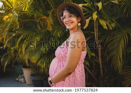 pregnant woman in a hat posing to the camera on a terrace with many plants on a nice sunny afternoon