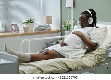 Pregnant woman with headphones sitting in hospital bed listening music while enjoying moment of relaxation and expecting baby in bright room with window blinds - Powered by Shutterstock