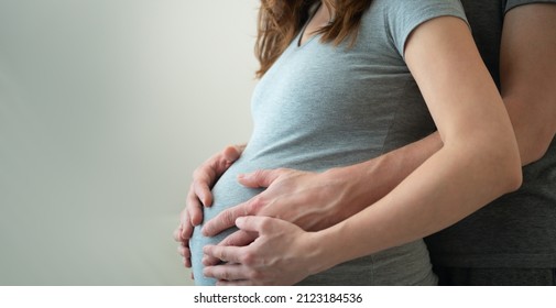 Pregnant Woman And Her Husband Holding Hand Together. Young Family Is Happy In Anticipation Of A Baby, Parenthood. Two Hands Holding Baby Inside A Pregnant Woman