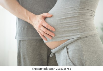 Pregnant Woman And Her Husband Holding Hand Together. Young Family Is Happy In Anticipation Of A Baby, Parenthood. Two Hands Holding Baby Inside A Pregnant Woman