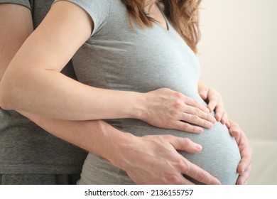 Pregnant Woman And Her Husband Holding Hand Together. Young Family Is Happy In Anticipation Of A Baby, Parenthood. Two Hands Holding Baby Inside A Pregnant Woman