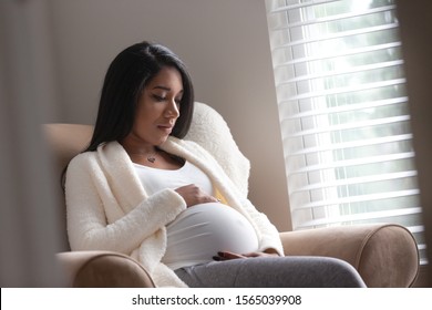 A pregnant woman holding her belly in a chair in deep thought