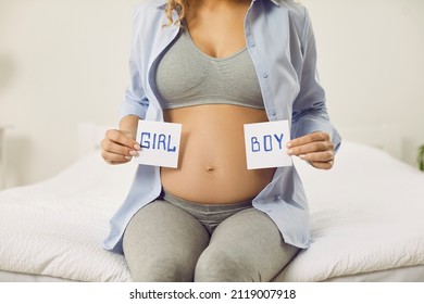 Pregnant Woman Holding Paper Cards With Inscriptions Boy And Girl Near Her Bare Belly. Cropped Image Of Unknown Woman Expecting Baby Sitting On Bed And Guessing Sex Of Baby. Gender Reveal Concept.