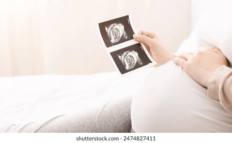 Pregnant woman holding ultrasound image of her baby and caressing her belly, lying in bed, empty space - Powered by Shutterstock