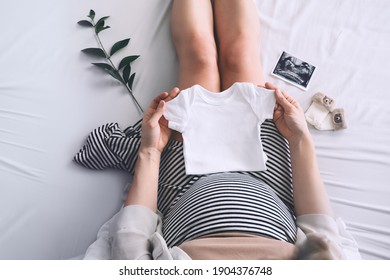 Pregnant woman holding white baby bodysuit preparing to child birth during pregnancy. Young mother with pregnant belly waiting of a baby.
