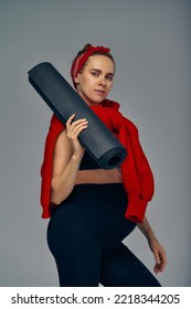 Pregnant Woman Holding Yoga Mat In Hands, Going For Sport While Awaiting Baby, Looking Smiling At Camera And Touching Her Belly, Standing Against Gray Wall.