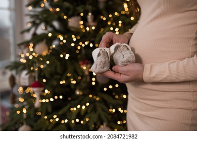 A Pregnant Woman Holds Knitted Shoes For A Baby In Her Hands On The Background Of A Christmas Tree, Pregnancy At Christmas, Christmas Surprise.