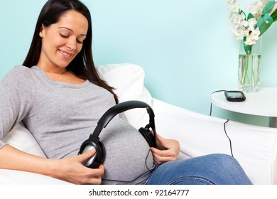 Pregnant woman at home sitting in an armchair playing music through headphones to her baby, it's suggested that this helps the baby to relax and that the baby may remember the music once they're born. - Powered by Shutterstock