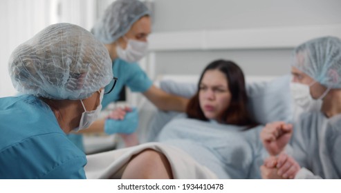 Pregnant woman in hospital giving birth with husband in mask holding her hand. Young lady pushing delivering baby in hospital with obstetrics team help - Shutterstock ID 1934410478