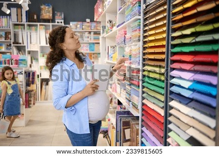 Pregnant woman, inspired artist painter holding sketchbook, standing by shelf with color paper sheets for design, pastel and watercolor painting in a creative art store. School stationery shop