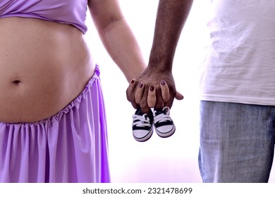 Pregnant woman, interracial couple holding hands holding baby booties. Close-up photo. - Shutterstock ID 2321478699