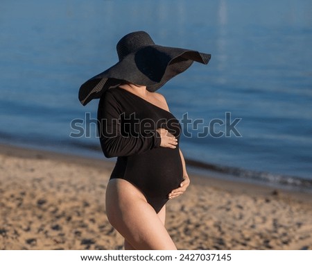 Pregnant woman in a large straw hat and black swimsuit posing on the beach.
