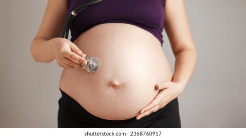 pregnant woman listening with a stethoscope to her big belly on a gray studio background concept of health examine - Shutterstock ID 2368760917