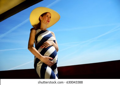 Pregnant woman, in long dress and large hat, holding belly, in front of blue sky with contrails