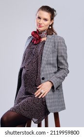 Pregnant Woman In A Loose Dress And Jacket Sitting On Chair On Grey Studio Background