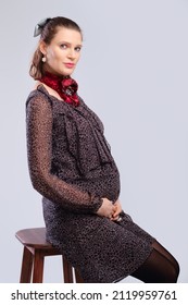 Pregnant Woman In A Loose Dress Sitting In Profile On Chair On Grey Studio Background