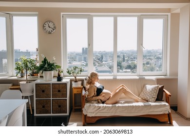 Pregnant Woman Lying On A Sofa In Soft Light By The Window At Home, Wearing A Cozy Cardigan.