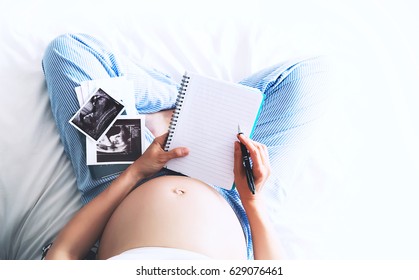 Pregnant woman makes notes in notebook and holding ultrasound image and medical documents at home. Pregnancy, parenthood, preparation and expectation concept. Close-up, copy space, indoors.