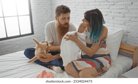 Pregnant woman and man sitting together on a bed in a bedroom, sharing a tender moment as they prepare for their baby by looking at a onesie and a stuffed toy. - Powered by Shutterstock