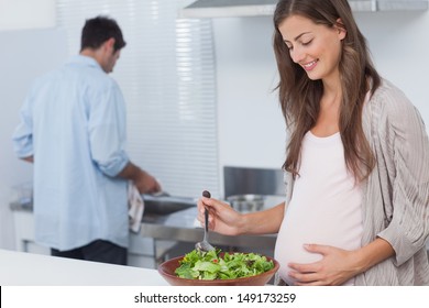 Pregnant woman mixing a salad in the kitchen while her husband is washing the dishes behind