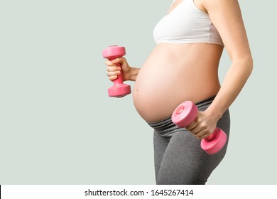 Pregnant Woman On A Gradient Green Background Holding Pink Weights, Wearing Yoga Pants And Sports Bra Exercising 