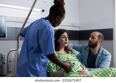 Pregnant woman with painful labor lying in hospital ward bed, having contractions. African american nurse and husband supporting future mother, holding hands for medical assistance - Powered by Shutterstock