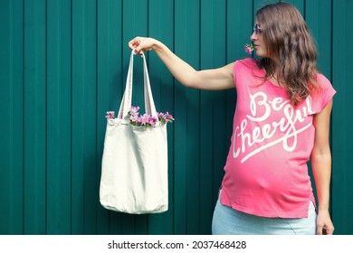 Pregnant Woman In Pink T-shirt With Be Cheerful Inscription Holding Blank Canvas Eco Shopping Bag With Flowers Inside On Green Background. Zero Waste Mock-up.