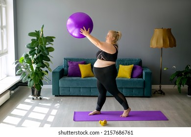 A Pregnant Woman With Plus-size Obesity Is Doing Fitness At Home In The Hallway Lifting The Fitball Up. A Woman In A Black Tracksuit Top And Leggings Is Engaged In Front Of The Sofa