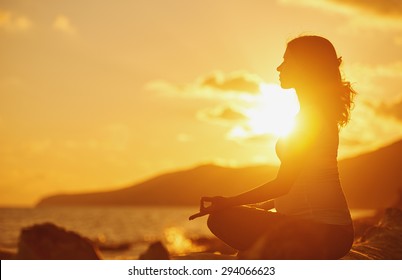 Pregnant woman practicing yoga, sitting in lotus position on a beach at sunset