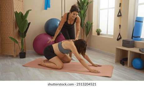 Pregnant woman practicing yoga with a smiling female trainer in an indoor gym setting with fitness equipment and natural light - Powered by Shutterstock