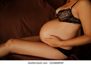 Pregnant woman pregnancy boudoir photoshoot in black lace underwear dark background. Profile body crop of lady touching expecting baby bump.