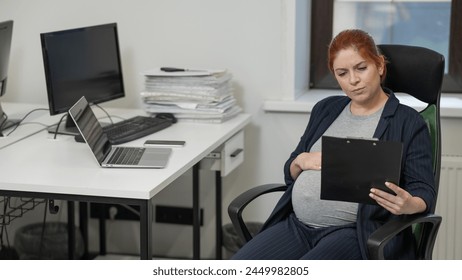 Pregnant woman reading documents on a paper tablet in the office.  - Powered by Shutterstock