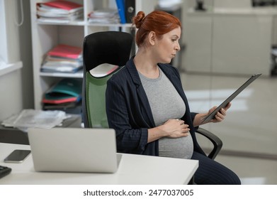 Pregnant woman reading documents on a paper tablet in the office.  - Powered by Shutterstock