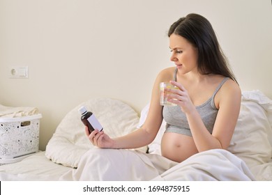 Pregnant Woman Reading Label On Bottle With Medicine, With Vitamins. Female Sitting At Home In Bed With Glass Of Water Medicine. Pregnancy, Health, Pharmaceuticals, Care And People.