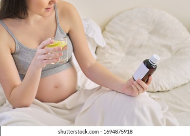 Pregnant Woman Reading Label On Bottle With Medicine, With Vitamins. Female Sitting At Home In Bed With Glass Of Water Medicine. Pregnancy, Health, Pharmaceuticals, Care And People.