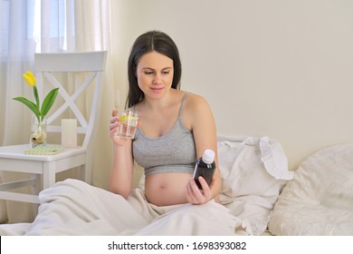 Pregnant Woman Reading Label On Bottle With Medicine, With Vitamins. Female Sitting At Home In Bed With Glass Of Water Medicine. Pregnancy, Health, Pharmaceuticals, Care And People.