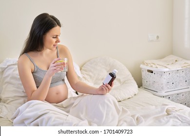 Pregnant Woman Reading Label On Bottle With Medicine, With Vitamins. Female Sitting At Home In Bed With Glass Of Water Medicine. Pregnancy, Health, Pharmaceuticals, Care And People.